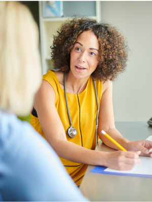 A gp in a yellow top with a stethoscope talking notes while talking to a patient 